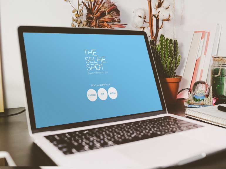 An open laptop displaying The Selfie Spot virtual photo booth blue start page, sitting on a desk with plants and a notebook with a pen.