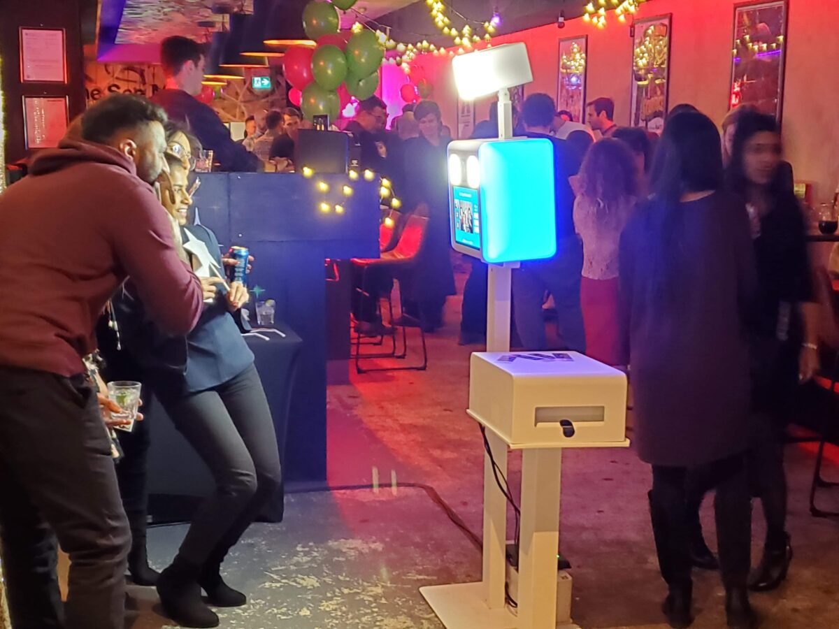 People posing and smiling in front of a photo booth at an evening event with drinks, twinkle lights and a bar area.