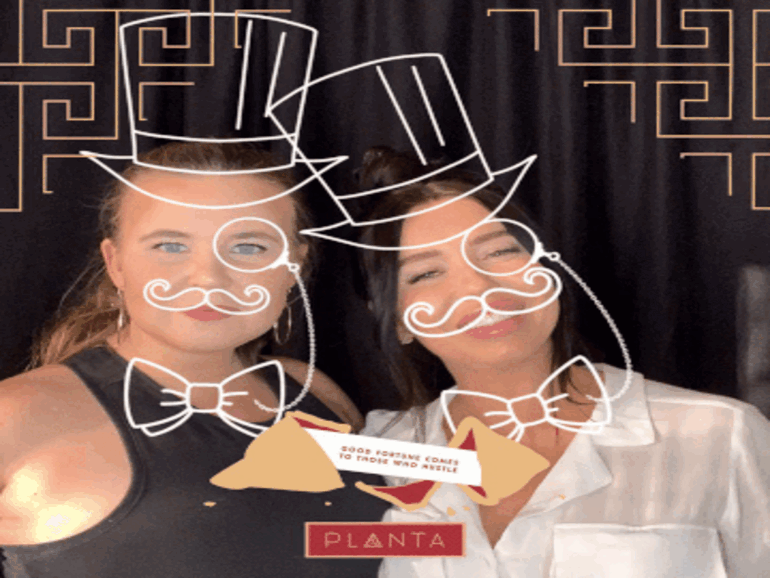 A GIF of two women smiling and posing in front of a social photo booth branded with a corporate logo, overlaid with top hats, moustaches and bow ties.