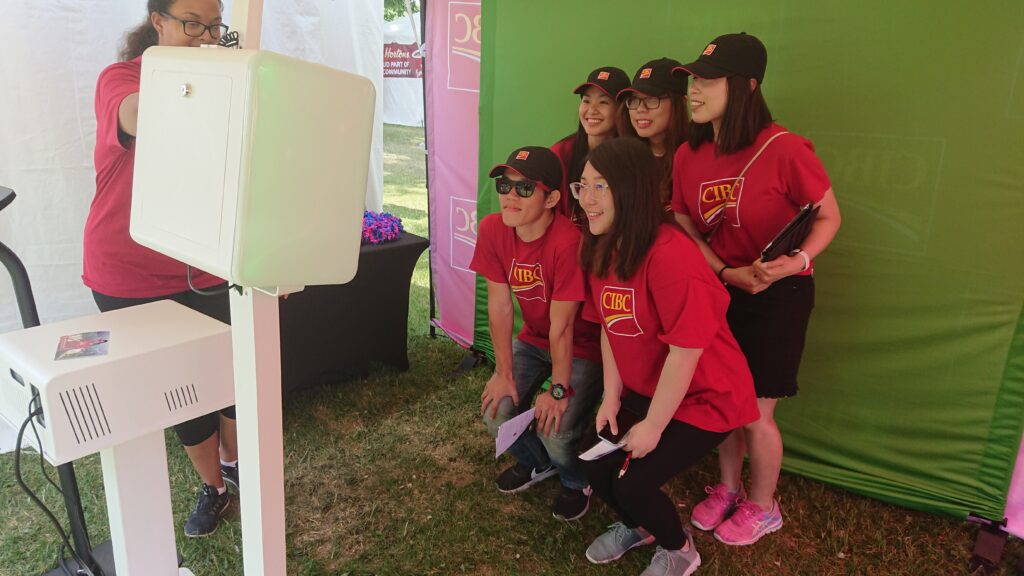 A white experiential marketing photo booth setup at an outdoor festival, showing a group of guests in matching red CIBC shirts posing in front of a physical green screen backdrop during a corporate brand activation