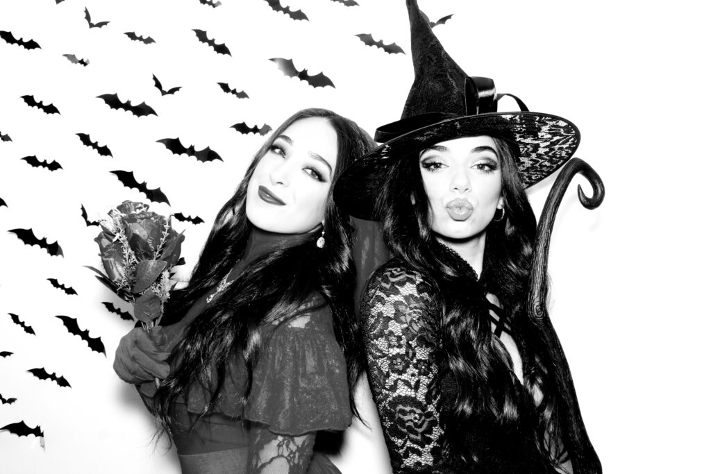 Two guests in witch costumes using a black and white glam photobooth and bat greenscreen for Halloween party ideas